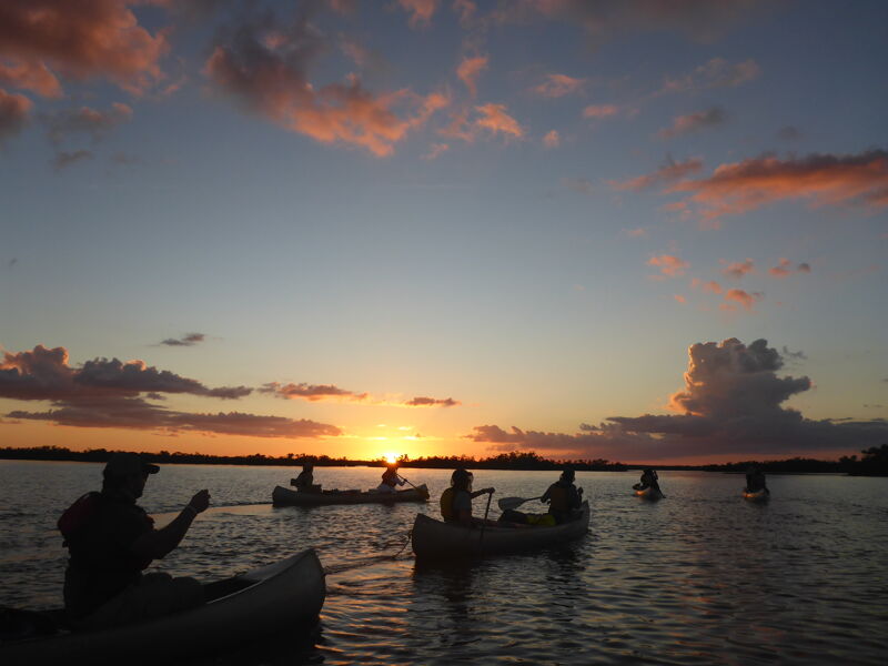 The image shows a serene scene at sunset with several canoes on a calm body of water. Silhouettes of people are visible in the canoes, paddling and enjoying the tranquil atmosphere. The sky is painted with hues of orange and pink, reflecting on the water's surface. The overall impression is one of peace and natural beauty.